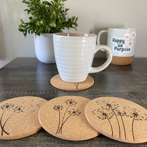 May include: Four cork coasters with a dandelion design. The coasters are round and have a natural cork finish. The dandelions are engraved into the cork and have a delicate, whimsical look.