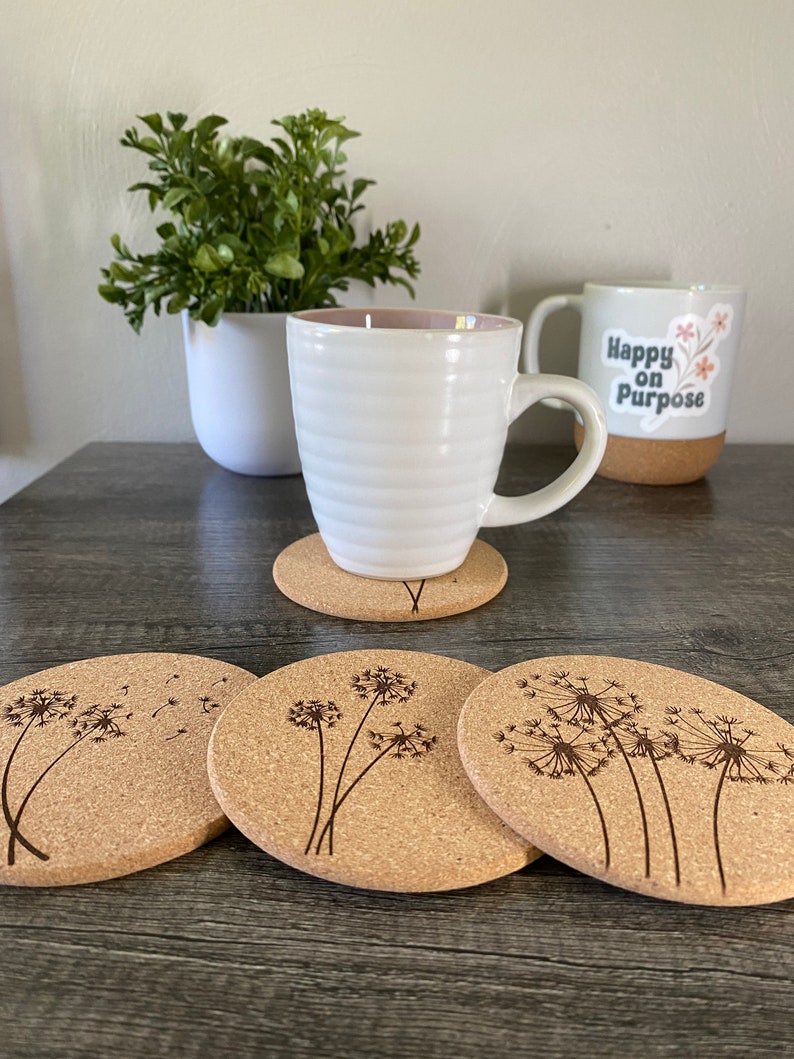 May include: Four cork coasters with a dandelion design. The coasters are round and have a natural cork finish. The dandelions are engraved into the cork and have a delicate, whimsical look.