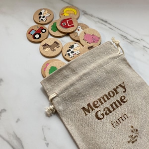 May include: A wooden farm-themed memory game with colorful illustrations of animals, a tractor, a barn, and a chick. The game pieces are in a beige drawstring bag with the text "Memory Game farm" printed on it.