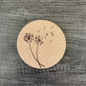May include: A round cork coaster with a brown dandelion design. The dandelion has seeds blowing in the wind.