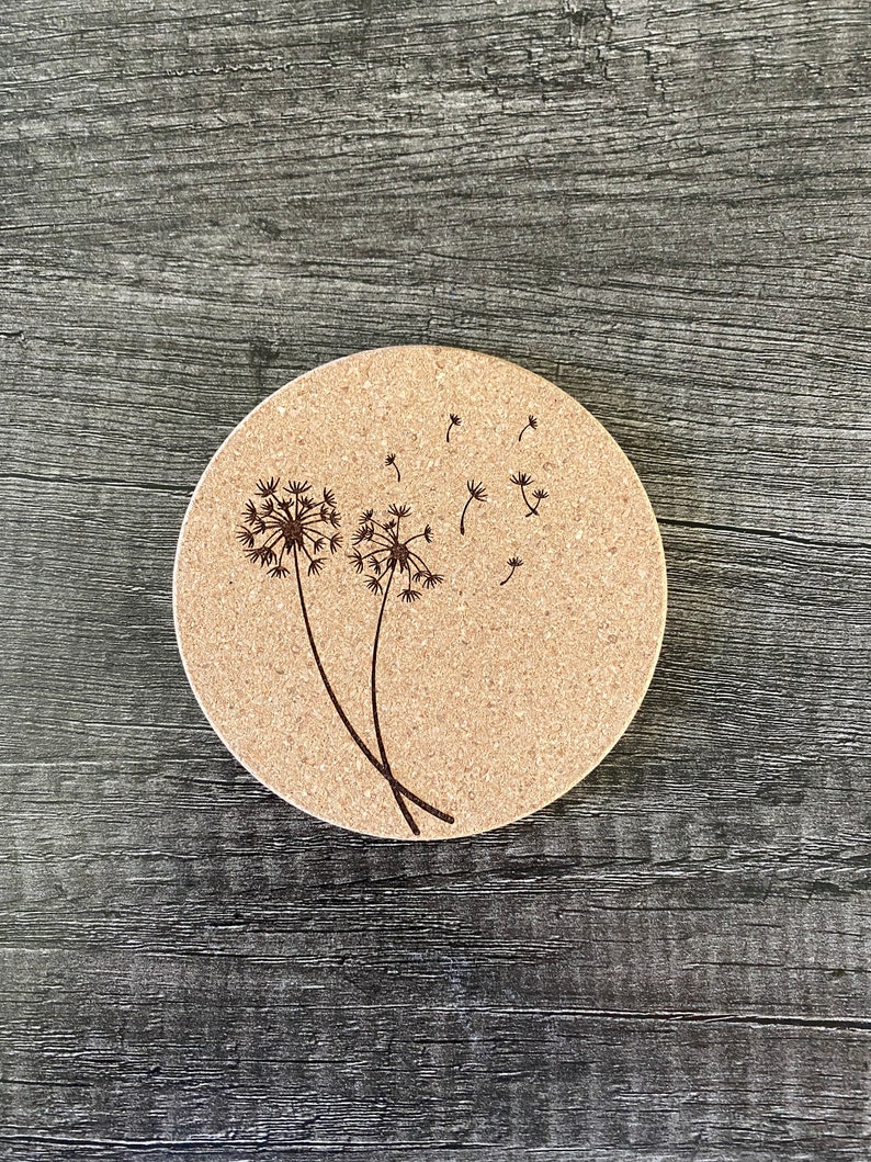 May include: A round cork coaster with a brown dandelion design. The dandelion has seeds blowing in the wind.
