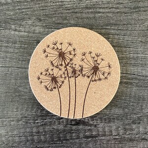 May include: A round cork coaster with a design of three dandelion flowers in brown on a beige background.