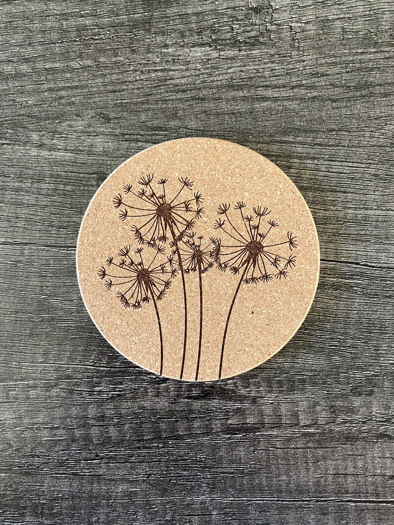 May include: A round cork coaster with a design of three dandelion flowers in brown on a beige background.