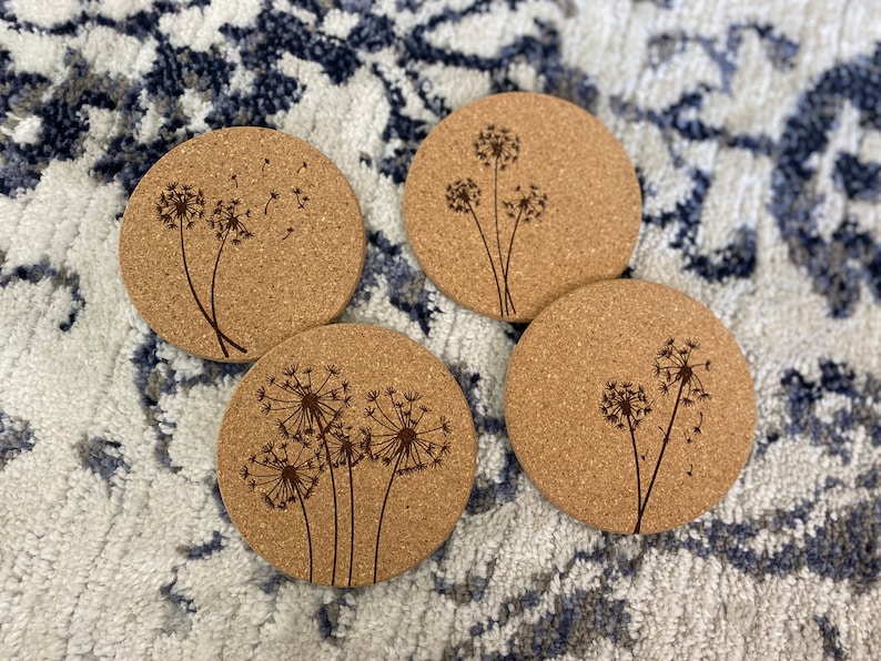 May include: Four round cork coasters with a laser-etched design of dandelions with seeds blowing in the wind.