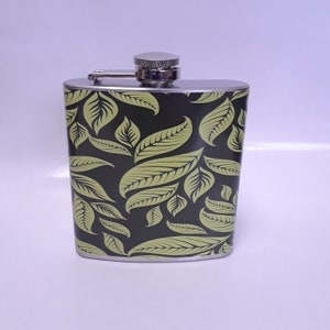 May include: A stainless steel flask with a black background and a repeating pattern of light green leaves. The flask has a silver cap and a hinged closure. The flask is designed to hold liquids and is a portable container.