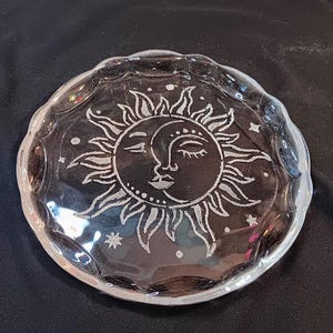 Puede incluir: Plato de vidrio transparente con borde festoneado. El plato presenta un diseño detallado de una cara de sol y luna con detalles de estrellas. El sol tiene rayos radiantes y la luna tiene una cara serena.