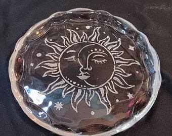 May include: Clear glass dish with a scalloped edge. The dish features a detailed design of a sun and moon face with star accents. The sun has radiating rays and the moon has a serene face.