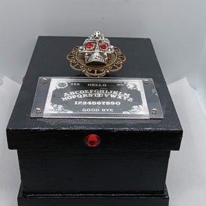 Peut inclure: Une boîte en bois noire avec un crâne décoratif et un tableau Ouija. Le crâne a des yeux en pierres rouges et est posé sur un filigrane en laiton. Le tableau Ouija affiche l'alphabet, les chiffres et les mots "Oui", "Bonjour", "Non" et "Au revoir".