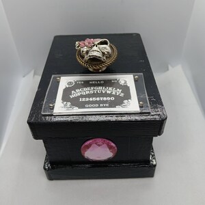 May include: A black rectangular box with a silver skull on top, decorated with pink flowers. The box has a planchette board with the text "YES", "HELLO", "NO", and "GOOD BYE". A large pink gem is on the front.