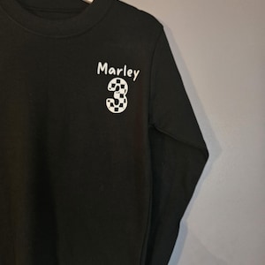 May include: Black long-sleeved t-shirt with a white chequered number "3" and the name "Marley" printed on the front.