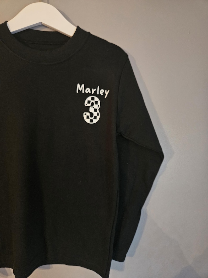 May include: Black long-sleeved t-shirt with a white chequered number "3" and the name "Marley" printed on the front.
