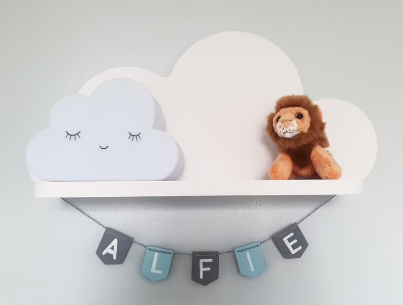 May include: White cloud shelf with a sleeping cloud nightlight and a stuffed lion. The shelf has a blue and gray banner that reads "Alfie" hanging below it.