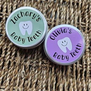 May include: Two small, round metal containers with lids, each featuring a cartoon tooth design. One lid is light green with the text "Zachary's Baby Teeth", and the other is purple with "Olivia's Baby Teeth". The containers sit on a woven, brown surface.