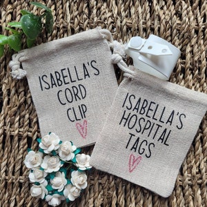 May include: Two small burlap bags with the text "Isabella's Cord Clip" and "Isabella's Hospital Tags" printed on them. Each bag has a pink heart design. A white cord clip is visible in the background.