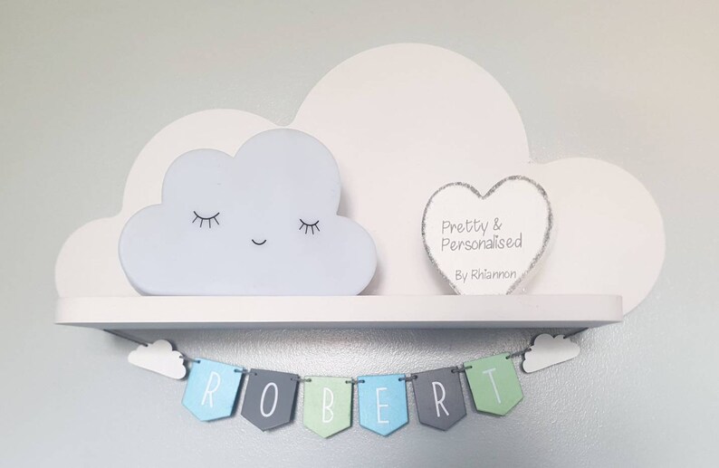 May include: A white wooden shelf with a cloud-shaped nightlight and a heart-shaped sign that reads "Pretty & Personalised By Rhiannon". A banner with the name "ROBERT" hangs below the shelf.