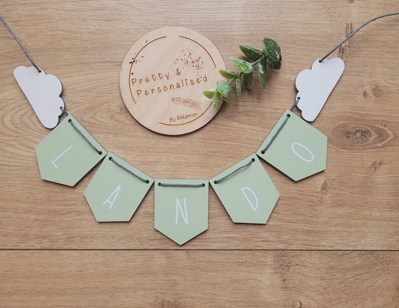 May include: A green and white wooden banner with the word "LANDO" spelled out in white letters. The banner is strung with gray string and has white cloud shapes on either end.