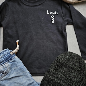May include: Black long-sleeved top with white text that says "Louis 1" on the front. The top is lying on a grey surface with a pair of blue jeans, a black beanie, and a black and white trainer.