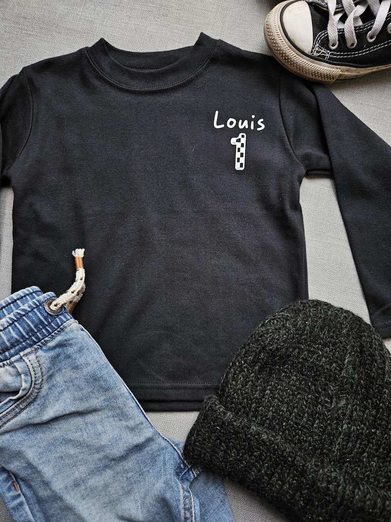 May include: Black long-sleeved top with white text that says "Louis 1" on the front. The top is lying on a grey surface with a pair of blue jeans, a black beanie, and a black and white trainer.