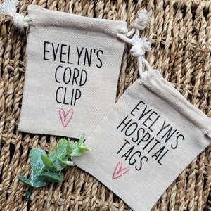 May include: Two small, beige fabric drawstring bags with red hearts and black text. The first bag says "Evelyn's Cord Clip" and the second bag says "Evelyn's Hospital Tags".