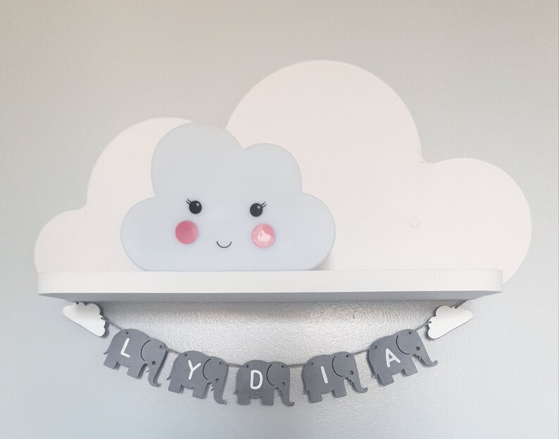 K&ouml;nnte beinhalten: Ein wei&szlig;es, wolkenf&ouml;rmiges Nachtlicht mit einem l&auml;chelnden Gesicht und rosa Wangen steht auf einem wei&szlig;en Regal. Unter dem Regal h&auml;ngt ein grauer Elefantenbanner mit dem Namen "LYDIA" in wei&szlig;en Buchstaben an einer wei&szlig;en Wolke.