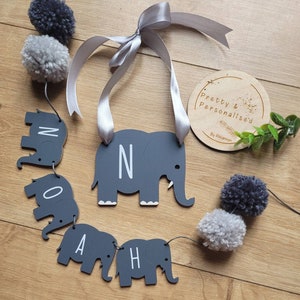 K&ouml;nnte beinhalten: Ein grauer Elefanten-Baby-Namen-Banner mit dem Namen NOAH in wei&szlig;en Buchstaben. Der Banner ist mit grauen und wei&szlig;en Pompons und einem grauen Satinband bespannt. Ein kleiner Holzkreis mit dem Text "Pretty & Personalised By Rhiannon" befindet sich im Hintergrund.