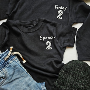 May include: Two black t-shirts with the names "Finley" and "Spencer" and the number "2" in a chequered pattern. The shirts are laid out on a white surface with a pair of blue jeans and a green beanie.