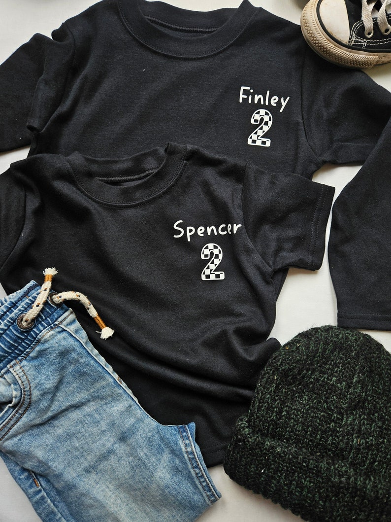 May include: Two black t-shirts with the names "Finley" and "Spencer" and the number "2" in a chequered pattern. The shirts are laid out on a white surface with a pair of blue jeans and a green beanie.