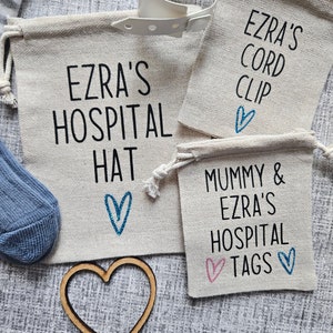 May include: Three small white canvas bags with blue and pink hearts and black text. The bags are labeled "Ezra's Hospital Hat", "Mummy & Ezra's Hospital Tags", and "Ezra's Cord Clip".
