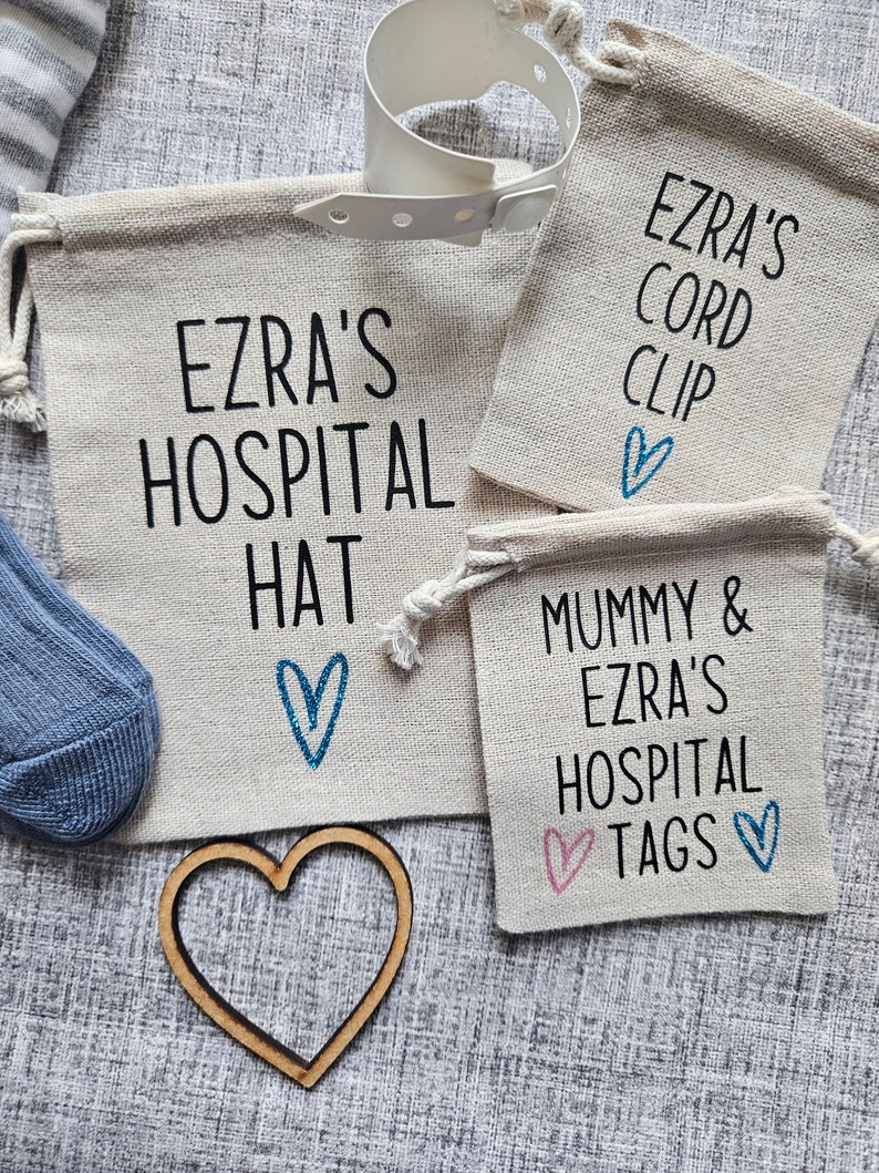 May include: Three small white canvas bags with blue and pink hearts and black text. The bags are labeled "Ezra's Hospital Hat", "Mummy & Ezra's Hospital Tags", and "Ezra's Cord Clip".