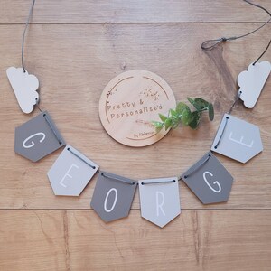 May include: A gray and white wooden banner with the name "GEORGE" spelled out in white letters. The banner is strung with gray twine and has two white cloud shapes at either end. A wooden circle with the text "Pretty & Personalised By Rhiannon" is in the center of the image.
