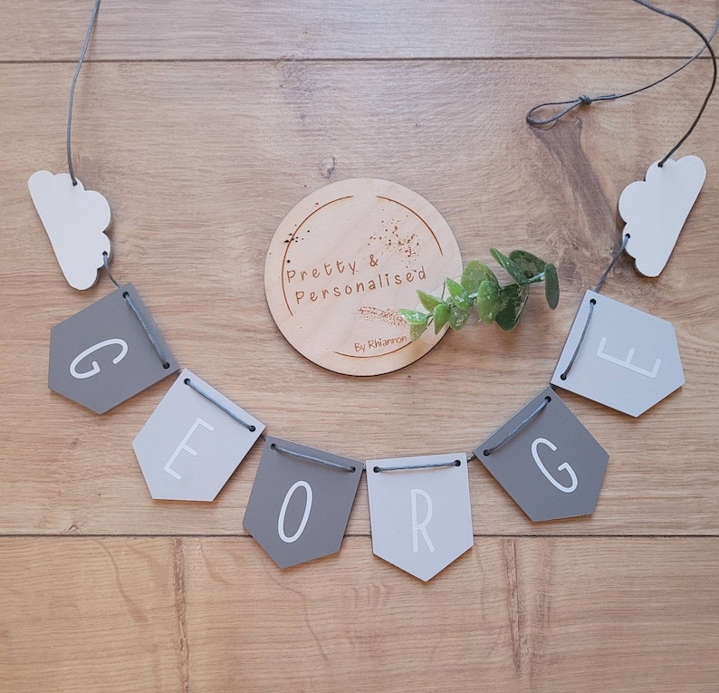 May include: A gray and white wooden banner with the name "GEORGE" spelled out in white letters. The banner is strung with gray twine and has two white cloud shapes at either end. A wooden circle with the text "Pretty & Personalised By Rhiannon" is in the center of the image.