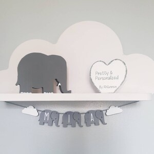K&ouml;nnte beinhalten: Ein wei&szlig;es, wolkenf&ouml;rmiges Regal mit einer grauen Elefantenfigur und einem grauen Herzen mit dem Text "Pretty & Personalised By Rhiannon". Unter dem Regal h&auml;ngt ein graues Elefantenbanner mit den Buchstaben "MIA".