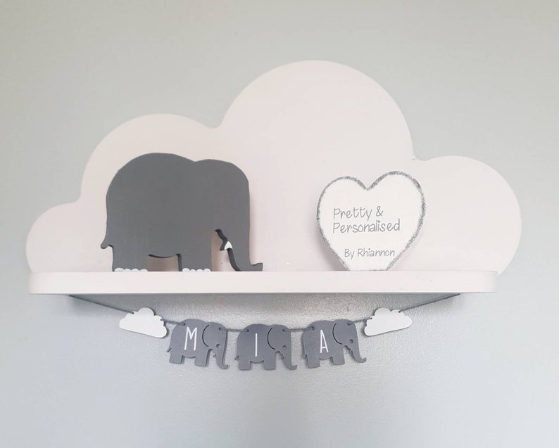 K&ouml;nnte beinhalten: Ein wei&szlig;es, wolkenf&ouml;rmiges Regal mit einer grauen Elefantenfigur und einem grauen Herzen mit dem Text "Pretty & Personalised By Rhiannon". Unter dem Regal h&auml;ngt ein graues Elefantenbanner mit den Buchstaben "MIA".