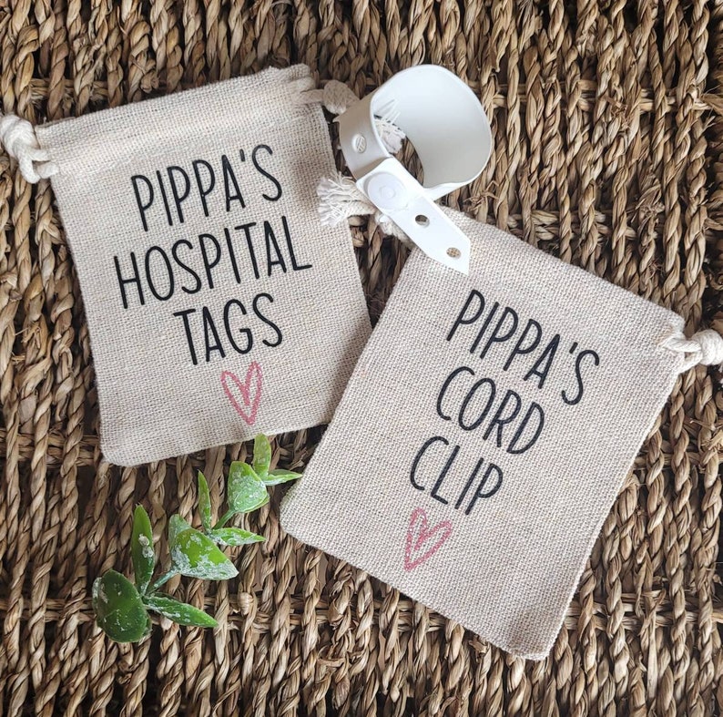 May include: Two small beige fabric bags with drawstring closures. The bags are decorated with black text that reads "Pippa's Hospital Tags" and "Pippa's Cord Clip". Each bag has a pink heart drawn on it.