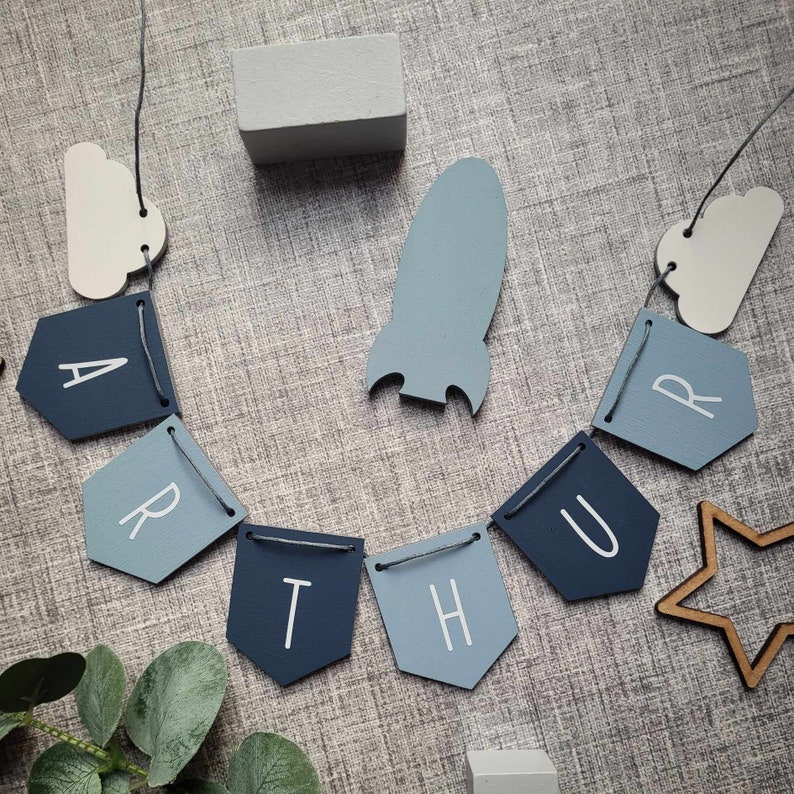 May include: A blue and white wooden banner with the letters "ARTHUR" spelled out. The banner is strung together with twine and has a rocket ship and cloud shapes hanging from it.