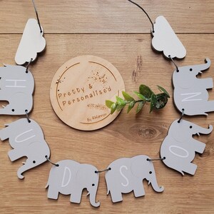 K&ouml;nnte beinhalten: Ein graues und wei&szlig;es Elefanten-Baby-Namensschild mit den Buchstaben "Hudson" ausgeschrieben. Das Schild ist mit wei&szlig;em Garn aufgef&auml;delt und hat an beiden Enden wei&szlig;e Wolken. Das Schild h&auml;ngt an einer Holzoberfl&auml;che.
