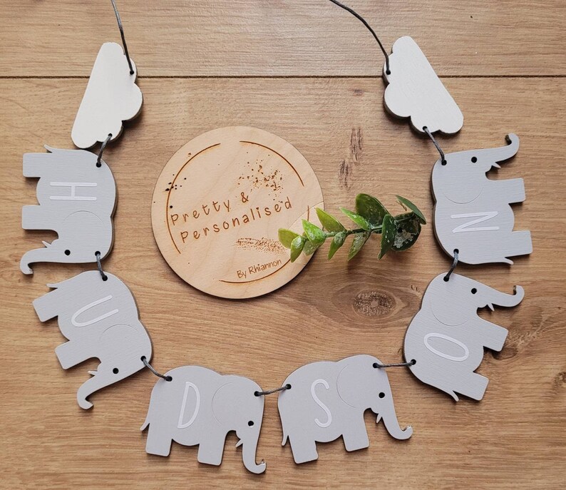 K&ouml;nnte beinhalten: Ein graues und wei&szlig;es Elefanten-Baby-Namensschild mit den Buchstaben "Hudson" ausgeschrieben. Das Schild ist mit wei&szlig;em Garn aufgef&auml;delt und hat an beiden Enden wei&szlig;e Wolken. Das Schild h&auml;ngt an einer Holzoberfl&auml;che.
