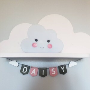 May include: A white cloud-shaped shelf with a smiling cloud-shaped light fixture. A banner with the name "DAISY" is hanging below the shelf.