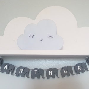 K&ouml;nnte beinhalten: Ein graues und wei&szlig;es Elefantenbanner, das den Namen "ARTHUR" buchstabiert, h&auml;ngt an einem wei&szlig;en Regal mit einem wei&szlig;en, wolkenf&ouml;rmigen Nachtlicht oben drauf.