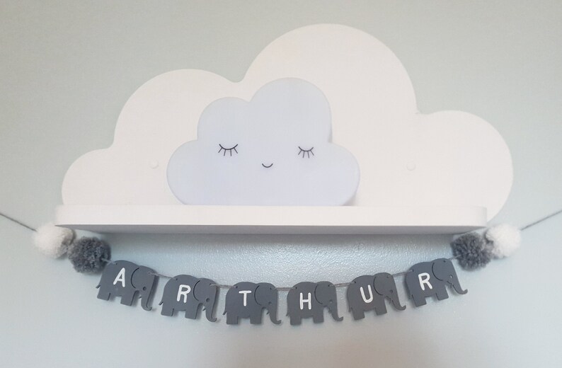 K&ouml;nnte beinhalten: Ein graues und wei&szlig;es Elefantenbanner, das den Namen "ARTHUR" buchstabiert, h&auml;ngt an einem wei&szlig;en Regal mit einem wei&szlig;en, wolkenf&ouml;rmigen Nachtlicht oben drauf.