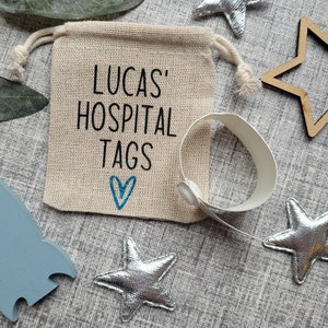 May include: A small burlap bag with a drawstring closure, printed with the text "Lucas' Hospital Tags" and a blue heart. The bag is filled with white hospital identification bracelets.