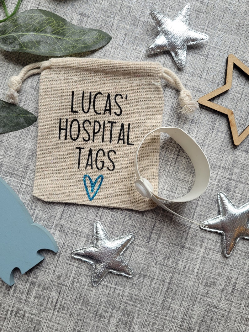 May include: A small burlap bag with a drawstring closure, printed with the text "Lucas' Hospital Tags" and a blue heart. The bag is filled with white hospital identification bracelets.