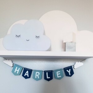 May include: A white shelf with a cloud-shaped nightlight and a wooden block. Below the shelf is a blue and white banner with the name "HARLEY" spelled out.