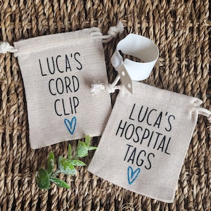 May include: Two small burlap bags with blue hearts and black text. The first bag says "Luca's Cord Clip" and the second bag says "Luca's Hospital Tags". A white hospital bracelet is visible in the background.