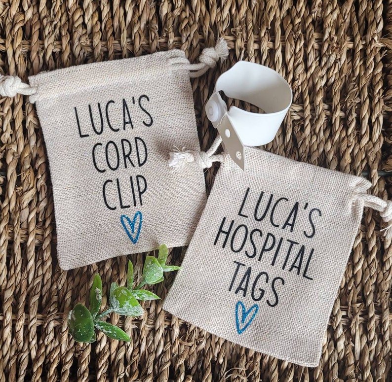 May include: Two small burlap bags with blue hearts and black text. The first bag says "Luca's Cord Clip" and the second bag says "Luca's Hospital Tags". A white hospital bracelet is visible in the background.
