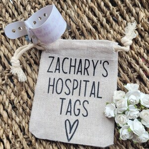 May include: A small, beige drawstring bag with the text "Zachary's Hospital Tags" and a heart printed on it. The bag is sitting on a woven surface.