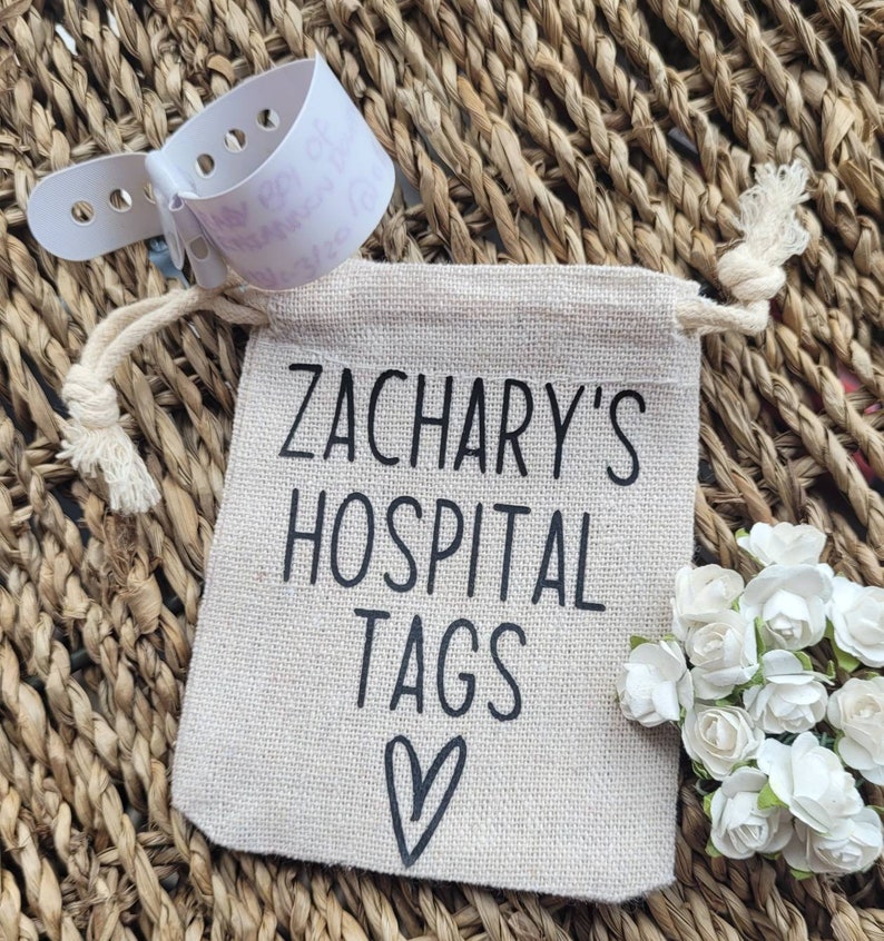 May include: A small, beige drawstring bag with the text "Zachary's Hospital Tags" and a heart printed on it. The bag is sitting on a woven surface.