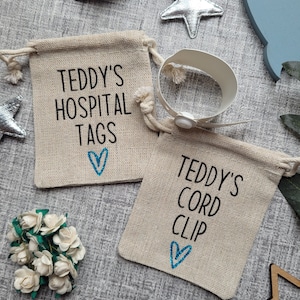 May include: Two small burlap bags with drawstring closures. The bags are personalized with the text "Teddy's Hospital Tags" and "Teddy's Cord Clip". Each bag has a blue heart embroidered on it. A white hospital bracelet with a snap closure is included.