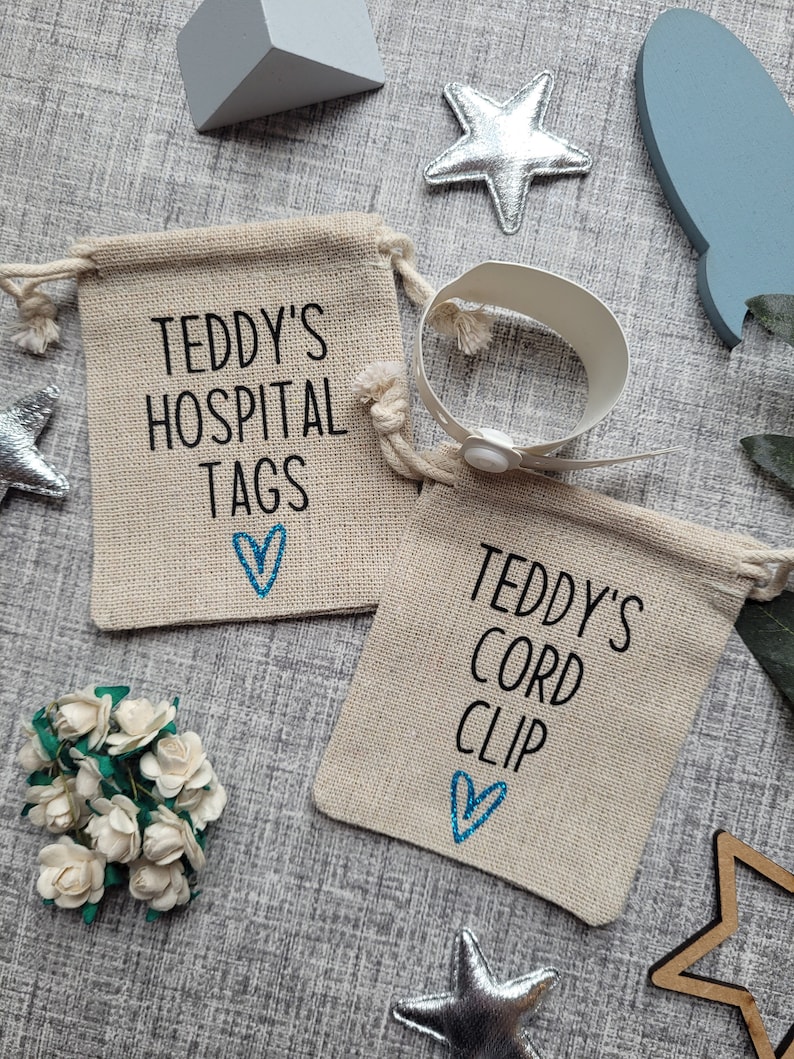 May include: Two small burlap bags with drawstring closures. The bags are personalized with the text "Teddy's Hospital Tags" and "Teddy's Cord Clip". Each bag has a blue heart embroidered on it. A white hospital bracelet with a snap closure is included.
