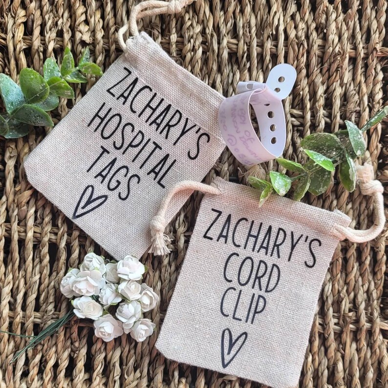 May include: Two small beige fabric bags with black text and a heart design. The bags are labeled "Zachary's Hospital Tags" and "Zachary's Cord Clip".